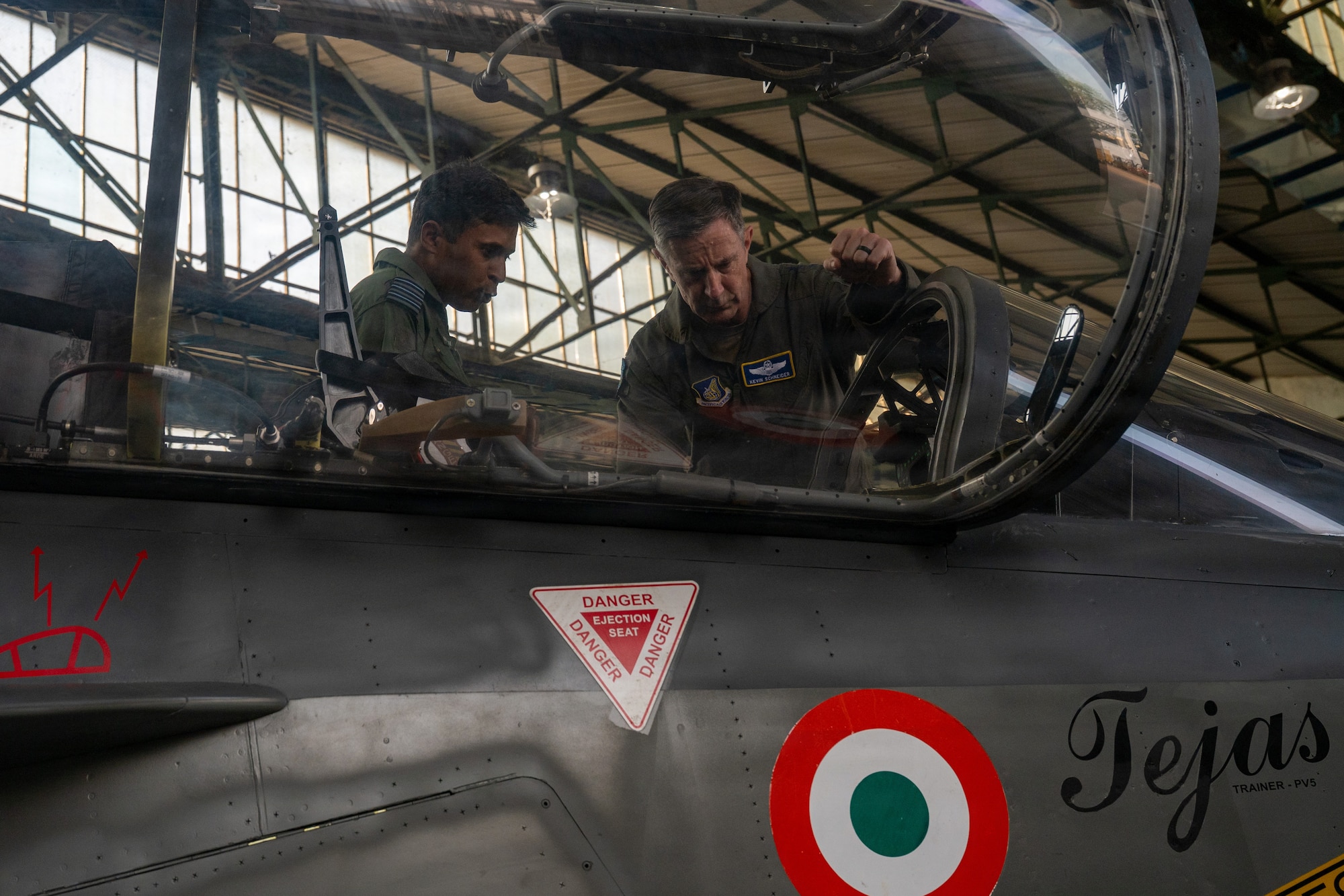 U.S. Air Force Gen. Kevin Schneider, in a flight suit, leans into the open cockpit of a Tejas fighter jet while speaking with an Indian Air Force pilot. The side of the aircraft has a visible Indian Air Force roundel and a 'DANGER EJECTION SEAT' warning sticker.