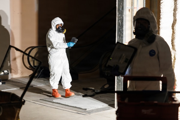 Two people in hazardous material suits monitor an empty room. One of the people holds a radiation detection device while the other person holds a tablet.