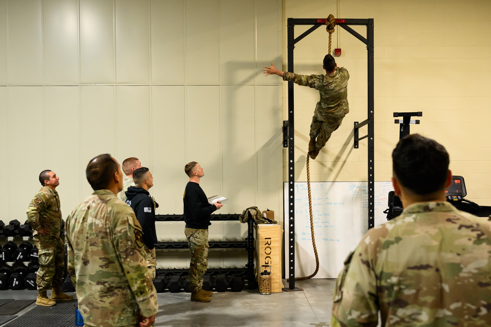 Airmen observe another Airman climbing up a rope inside a gym.