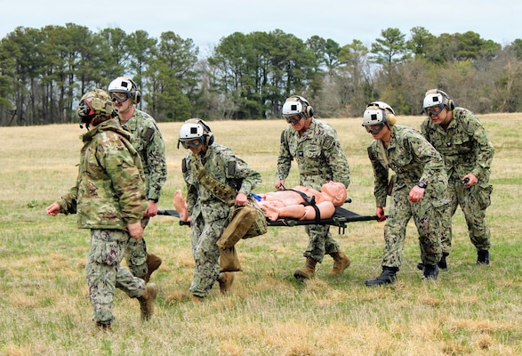 Capt. Ken Radford, center background, Commanding Officer of Navy Medicine Readiness and Training Command (NMRTC) Patuxent River, transports a simulated casualty on a litter alongside hospital corpsmen during a Tactical Combat Casualty Care (TCCC) tactical evacuation care (TACEVAC) drill at Naval Air Station Patuxent River, Maryland. The practical field scenario involved coordinating with an MH-60S Sea Hawk helicopter from the NAS Patuxent River Search and Rescue (SAR) "SAR Dogs" aircrew, testing patient transport protocols, signal smoke marking, and live casualty hand-off under simulated field conditions. The evolution was held as part of the command's "Warrior Wednesday" initiative to reinforce operational readiness and expeditionary trauma skills.