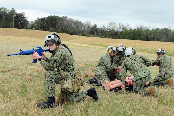 U.S. Navy Hospital Corpsman 3rd Class Timothy McWhorter, left, pulls security with a simulated training rifle while fellow corpsmen render point-of-injury medical care and prepare a mannequin for litter transport during a Tactical Combat Casualty Care (TCCC) evolution at Naval Air Station Patuxent River, Maryland. The practical scenario tested Tactical Field Care (TFC) principles—combining perimeter defense, MARCH algorithm trauma interventions, and litter team packaging. The drill was conducted as part of Navy Medicine Readiness and Training Command (NMRTC) Patuxent River’s "Warrior Wednesday" initiative to build expeditionary muscle memory and operational readiness across the command