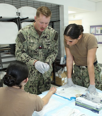 U.S. Navy Hospital Corpsman 1st Class Gregory Evanko, center, instructs Hospital Apprentice Elisa Medina Moya, right, as she prepares to administer intravenous (IV) access on Hospital Apprentice Kaitlyn Lewis during a hands-on Tactical Combat Casualty Care (TCCC) skill station at Navy Medicine Readiness and Training Command (NMRTC) Patuxent River, Maryland. The practical clinical drill focused on fluid resuscitation and circulation protocols under the MARCH algorithm. The evolution was conducted as part of NMRTC Patuxent River’s "Warrior Wednesday" initiative, designed to refine critical trauma techniques and maintain operational readiness across all medical personnel.