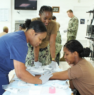U.S. Navy Hospital Corpsman 1st Class Erlande Alcius, center, Leading Petty Officer of Staff Education and Training, mentors Hospital Apprentice Christoria Bellamy, left, as she practices intravenous (IV) insertion techniques on Hospitalman Kristina Leggett during a Tactical Combat Casualty Care (TCCC) skill station. Alcius oversees logistics and curriculum development for command-wide training. The practical evolution was conducted as part of Navy Medicine Readiness and Training Command (NMRTC) Patuxent River’s "Warrior Wednesday" initiative, designed to refine critical clinical skills and maintain operational readiness across the command.