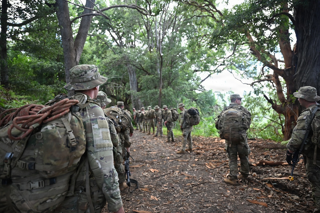 A dozen people in camouflage military uniforms walk down a dirt path in the jungle.