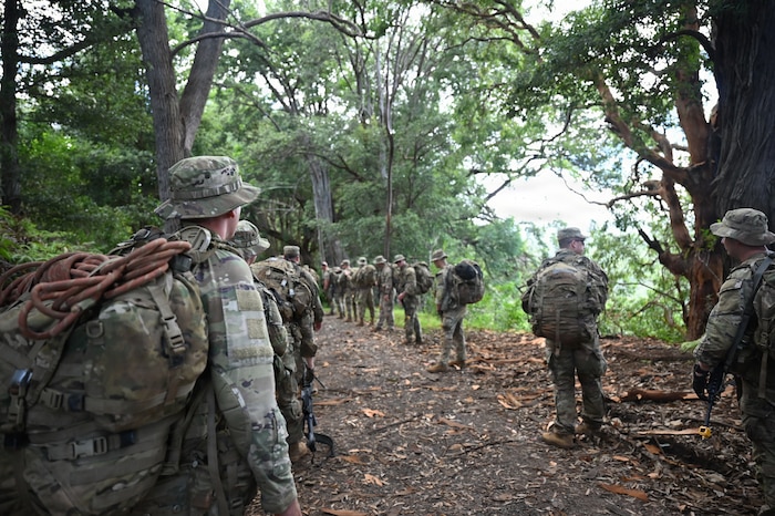 A dozen people in camouflage military uniforms walk down a dirt path in the jungle.