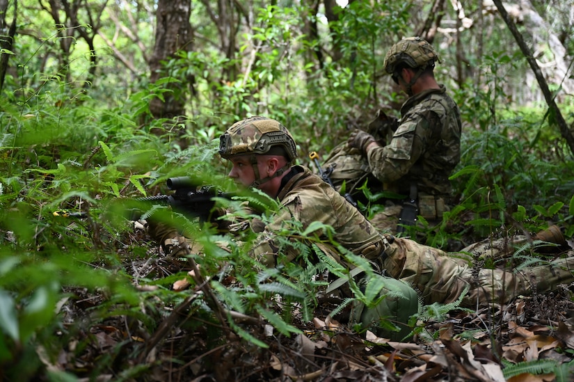 A man in a camouflage military uniform and helmet lies on the ground in the jungle aiming a rifle; there is another man in similar attire checking his equipment in the background.
