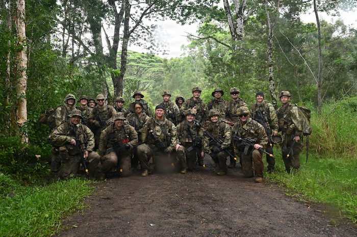 Twenty people in camouflage military uniforms and tactical gear pose for a photo on a dirt path in the jungle.