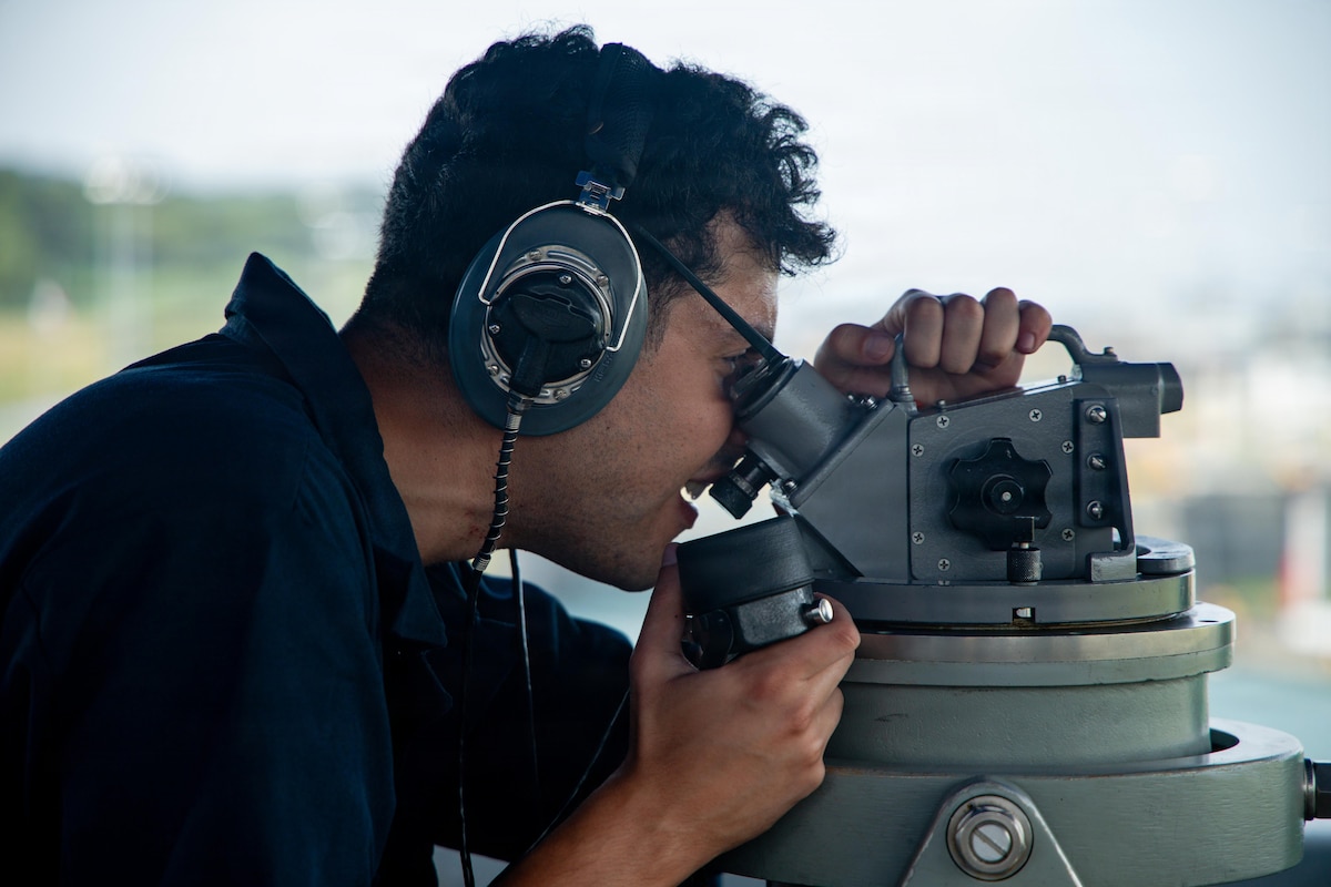 A man in a military uniform checks navigational equipment on a ship.