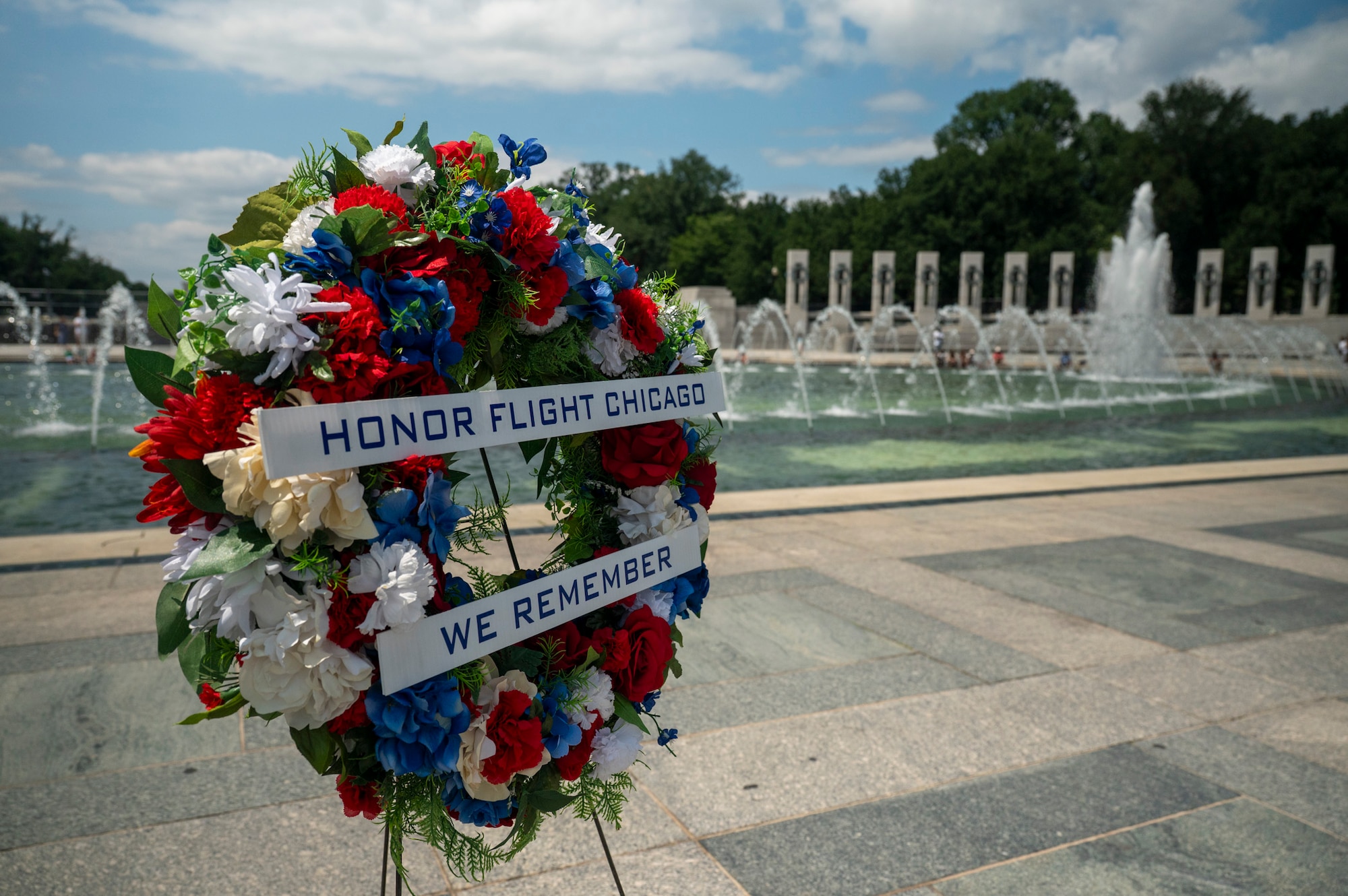 A wreath with red, white and blue flowers saying "Honor Flight Chicago, We remember" is displayed in front of the WWII memorial