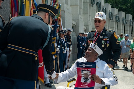 WWII veteran wearing his uniform shakes hands with members of the Joint Armed Forces Color guard in front of the WWII memorial