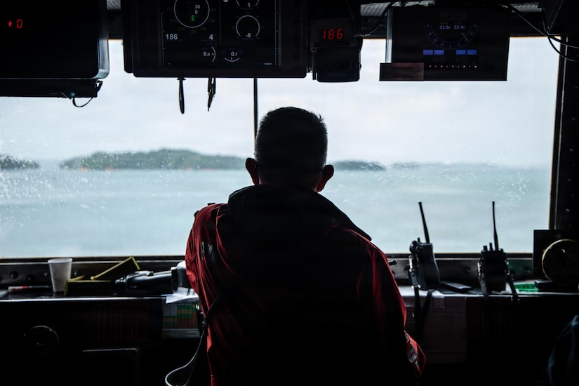 A man in a red jacket looks out a window in the dark bridge of a ship or building.
