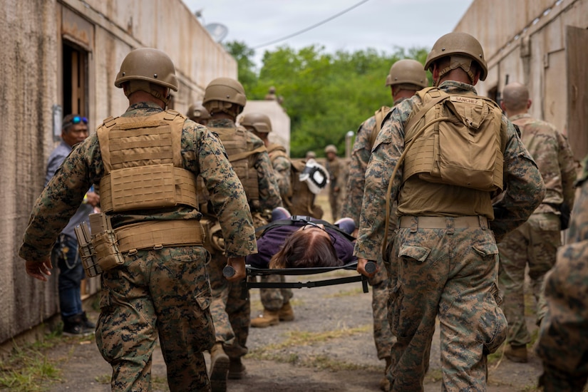Four people in camouflage military uniforms and combat gear carry a litter with a