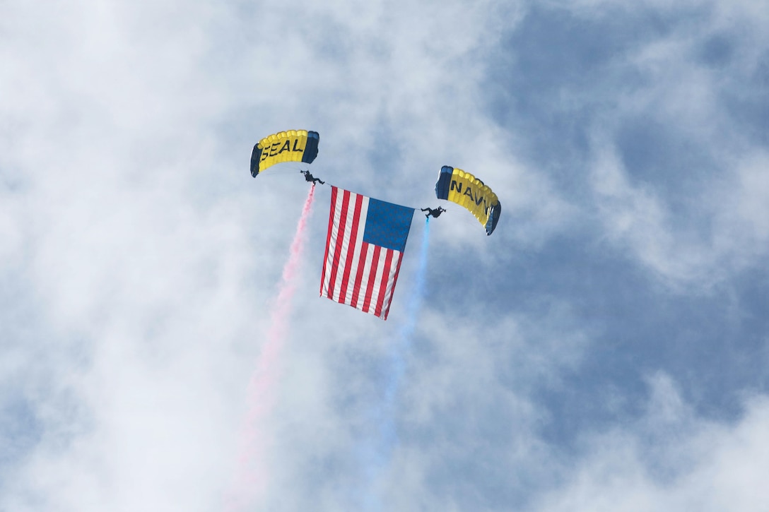 Two sailors, one spraying red smoke and the other spraying blue, display an American flag in a blue sky while descending with parachutes.