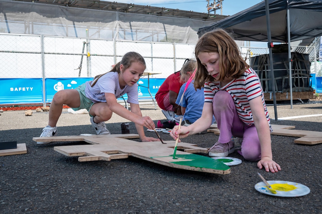 A military child kneels while another squats to paint a wooden aircraft on the ground during the day.