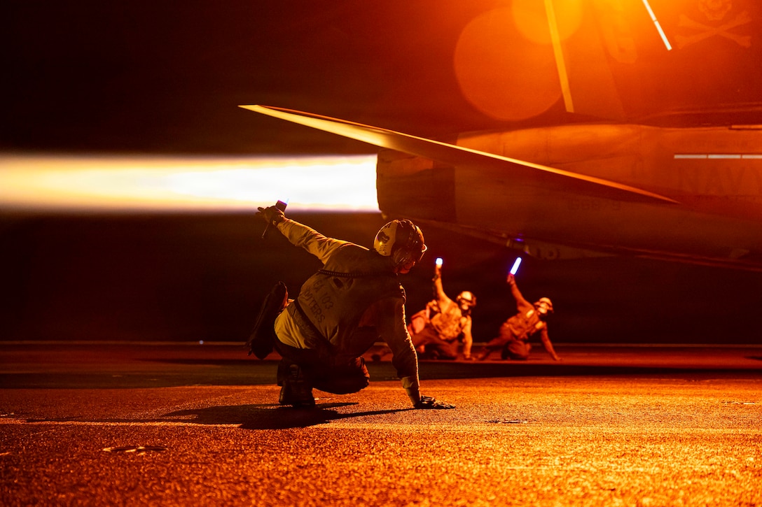 Three sailors lean on the deck of a ship with one hand while raising the other as a partially visible aircraft prepares to take off in the dark, leaving behind a bright streak.
