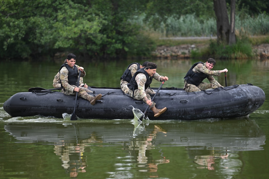 Five people in camouflage uniforms and black vests use oars to row a small inflatable boat in Army green water. Trees are in the background.