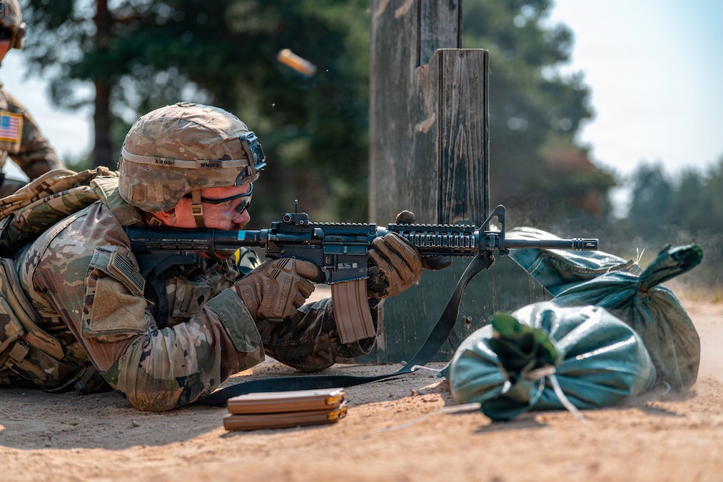 A person in a camouflage uniform and helmet fires an assault rifle while on the ground behind sandbags. A shell casing is in the air.