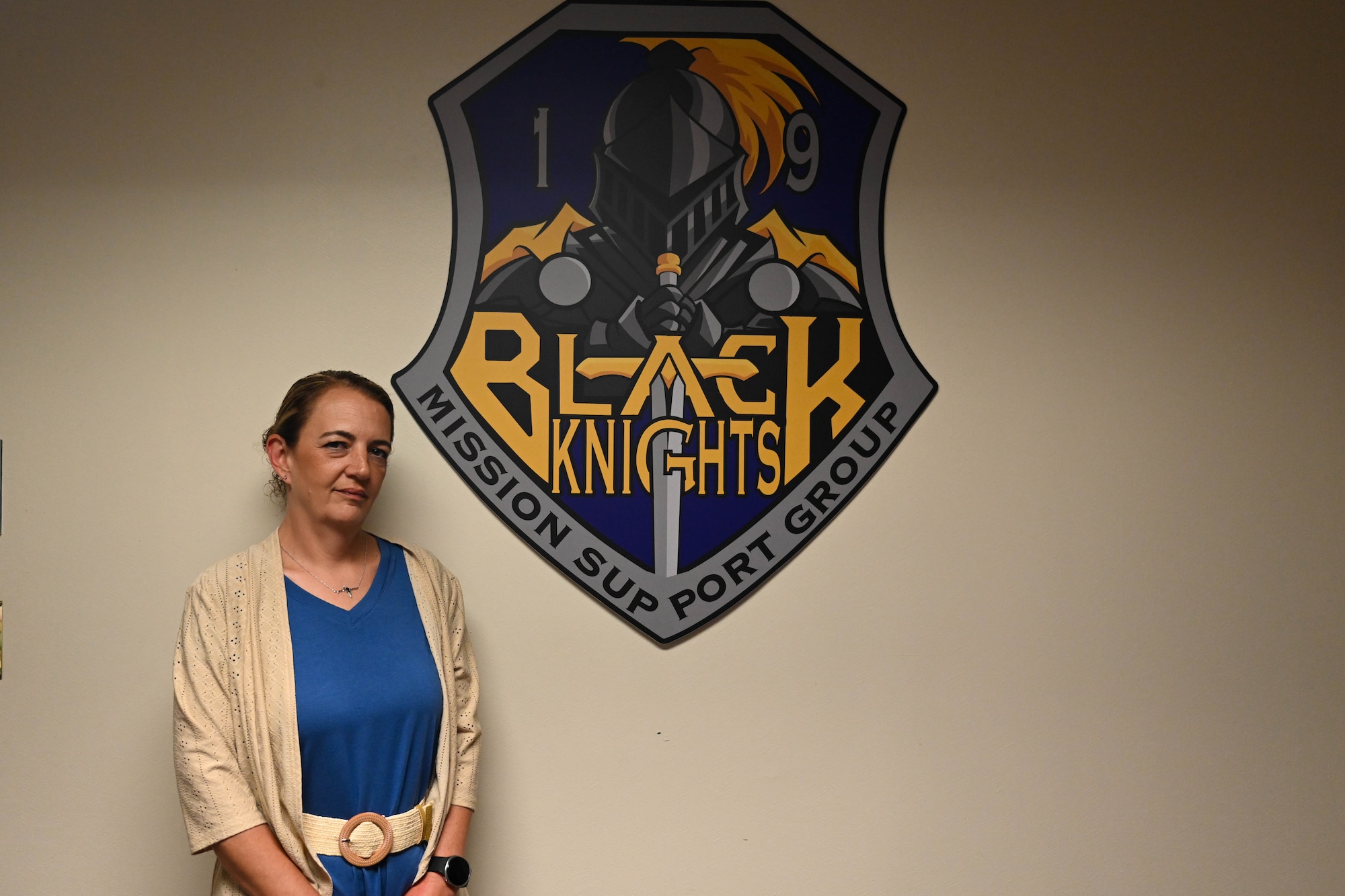 Woman stands in front of 19th Mission Support Group wall emblem.