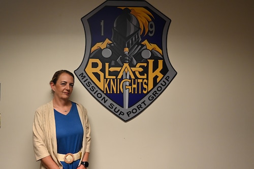 Woman stands in front of 19th Mission Support Group wall emblem.
