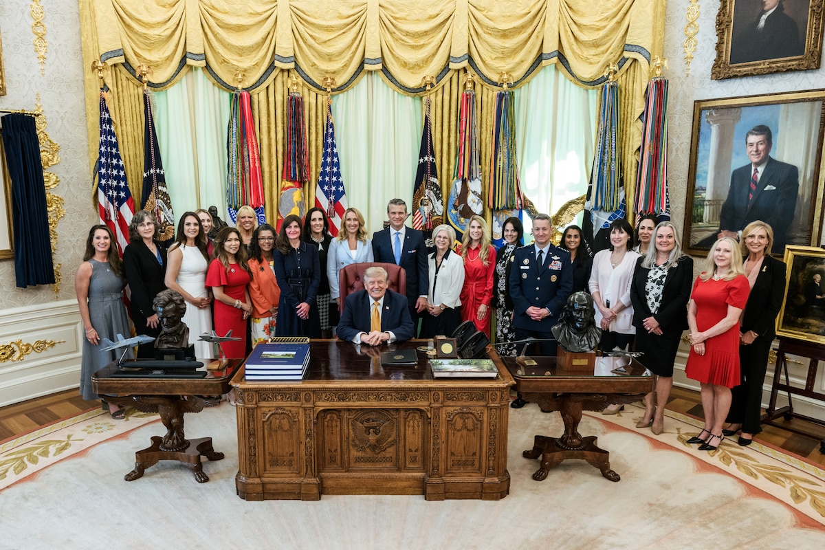 A man in business attire sits at a large wooden desk inside an office, surrounded by roughly two dozen men and women.