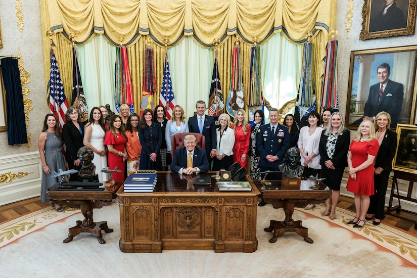 A man in business attire sits at a large wooden desk inside an office, surrounded by roughly two dozen men and women.