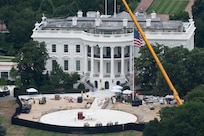 An aerial view of a large white building during daytime, with a large American flag and construction crane in front of it, along with a circular construction zone.