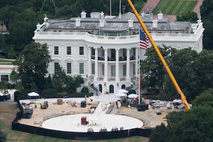 An aerial view of a large white building during daytime, with a large American flag and construction crane in front of it, along with a circular construction zone.