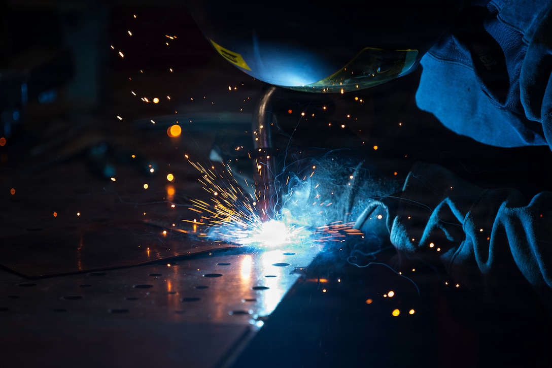 Sparks fly as an airman wearing protective gear welds medal in a dark room.