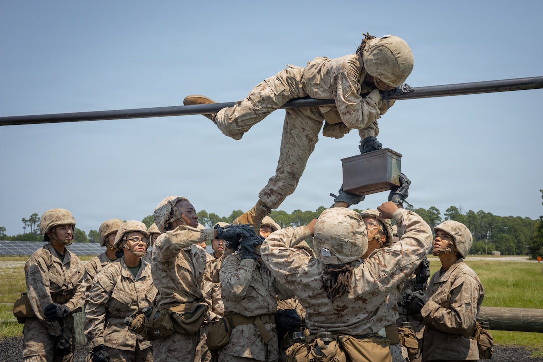 A group of Marine Corps recruits help a fellow recruit hanging on a horizontal line while carrying an ammo can against a blue sky.