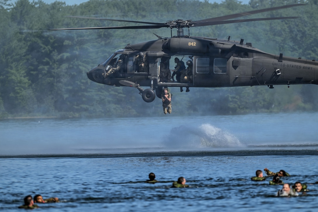 Soldiers swim in a body of water as another soldier jumps from a hovering helicopter during day.