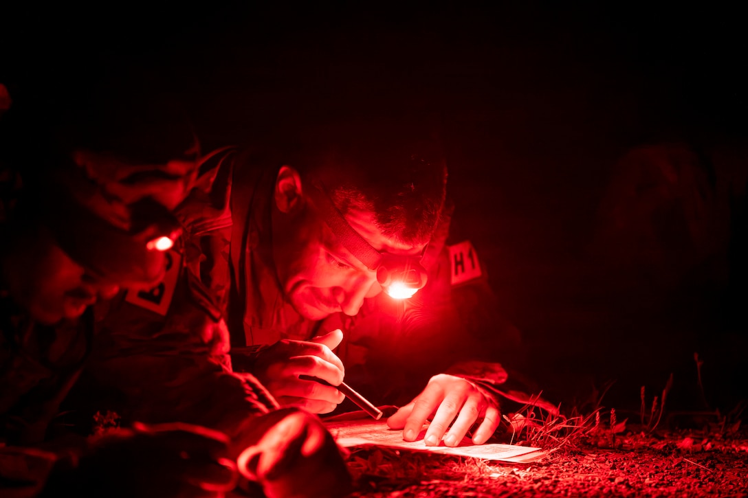 Two soldiers lying on the ground use red-lens headlamps while plotting points on a paper in the dark.