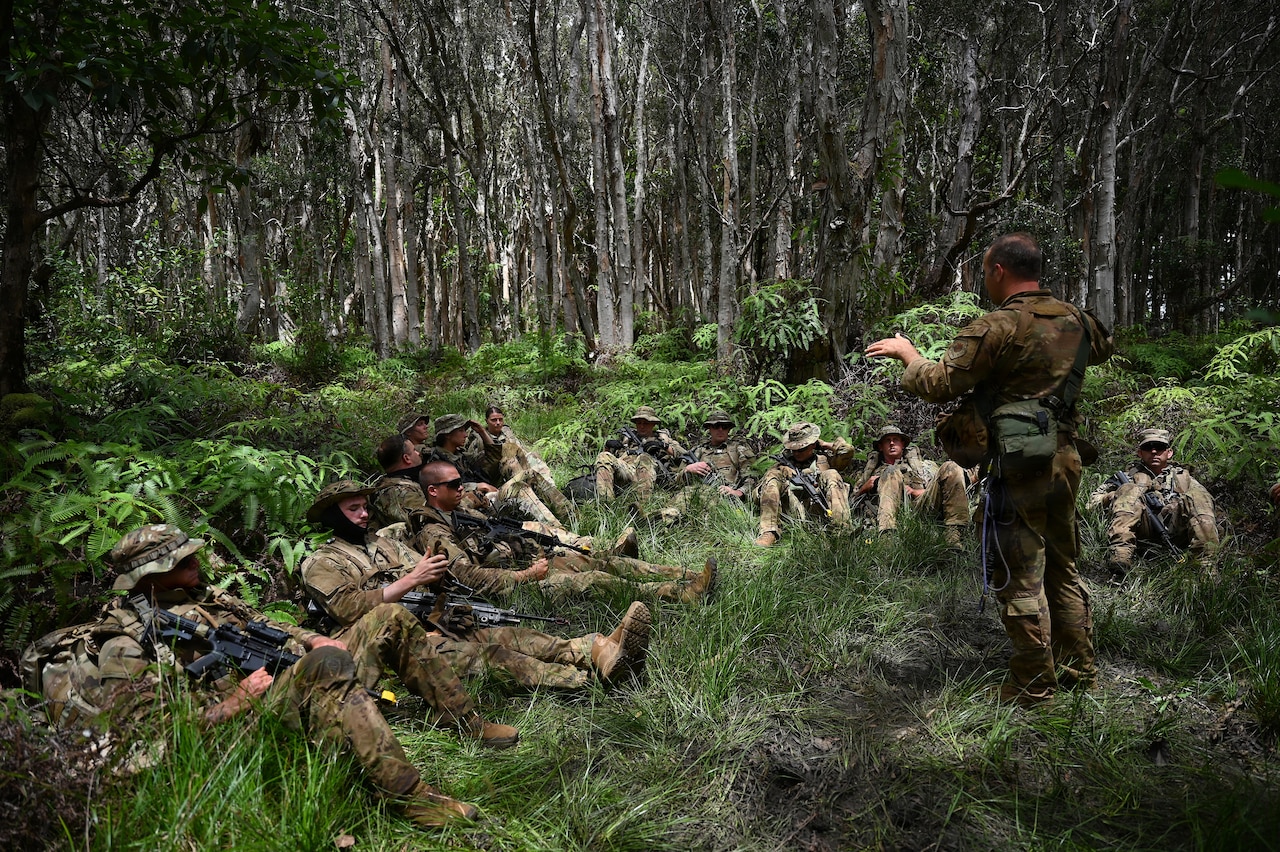 A man in a camouflage military uniform and tactical gear speaks to a dozen people in similar attire sitting in the jungle.