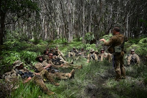 A man in a camouflage military uniform and tactical gear speaks to a dozen people in similar attire sitting in the jungle.