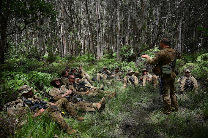 A man in a camouflage military uniform and tactical gear speaks to a dozen people in similar attire sitting in the jungle.