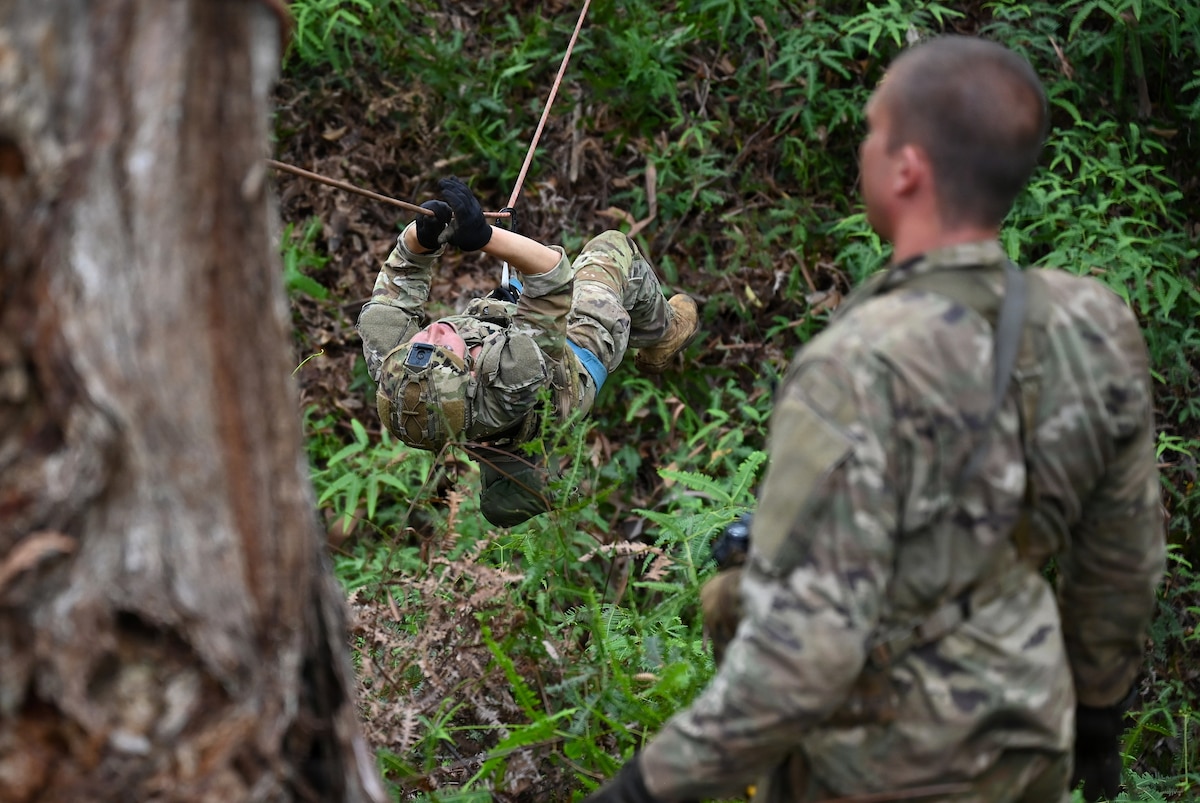 A person in a camouflage military uniform uses a rope to cross an area in the jungle as another person in similar attire observes.