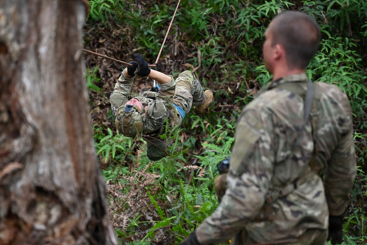 A person in a camouflage military uniform uses a rope to cross an area in the jungle as another person in similar attire observes.