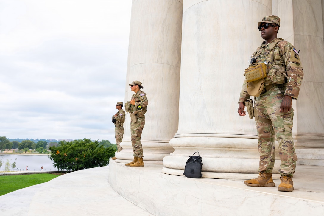 Three people in camouflage uniforms stand guard at the top of steps at a memorial. A tidal basin is in the background.