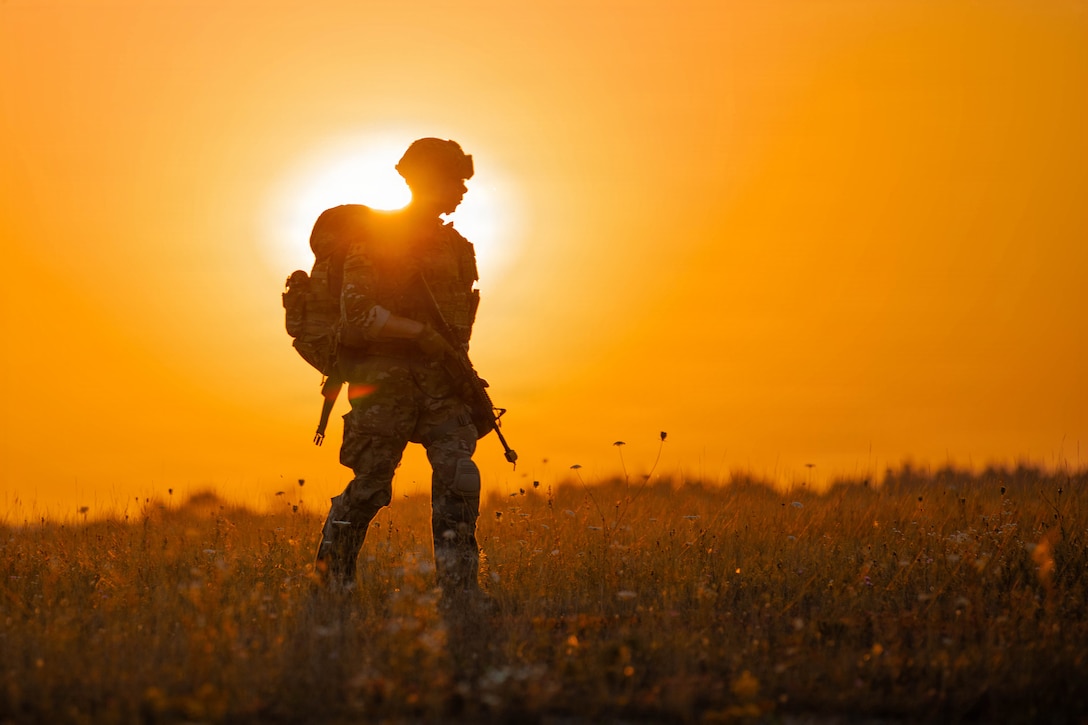 A person in camouflage carries a rifle while walking through a field. The sun and an orange-colored sky are in the background.