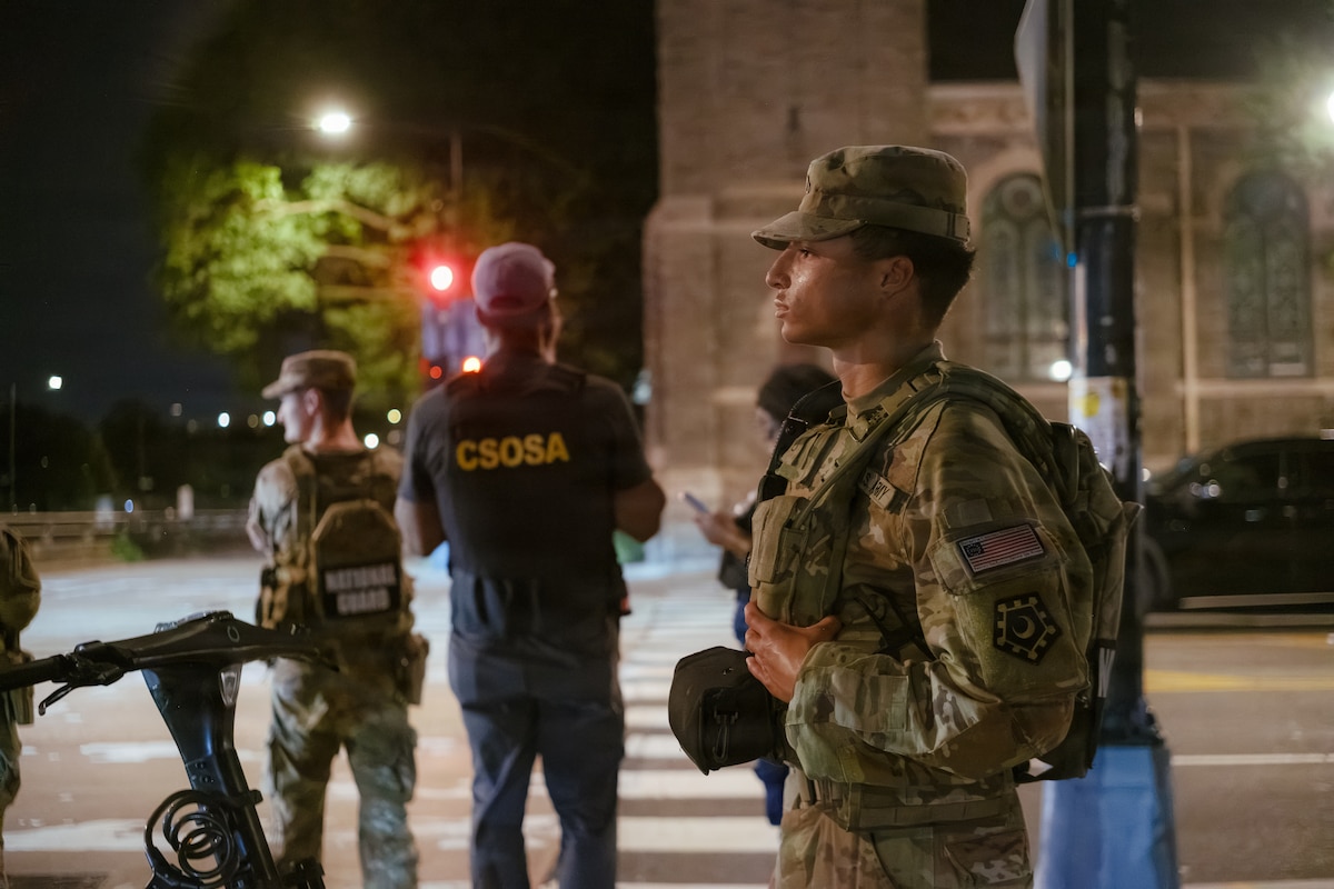 A side view of a soldier looking out while standing on a dark street corner illuminated by a light.