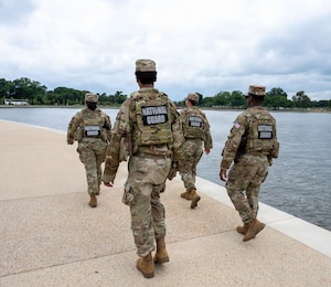 Four soldiers walk next to a small body of water near a monument during the day.