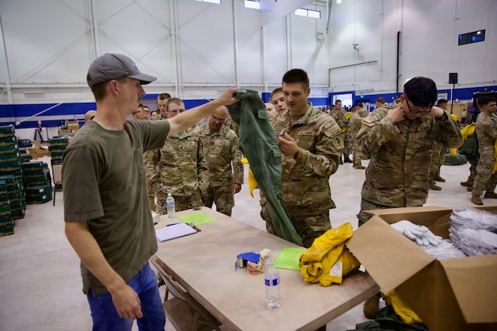 A group of people in military camouflage uniforms stand around inside a warehouse as they wait to be issued firefighter gear by a man in casual attire.