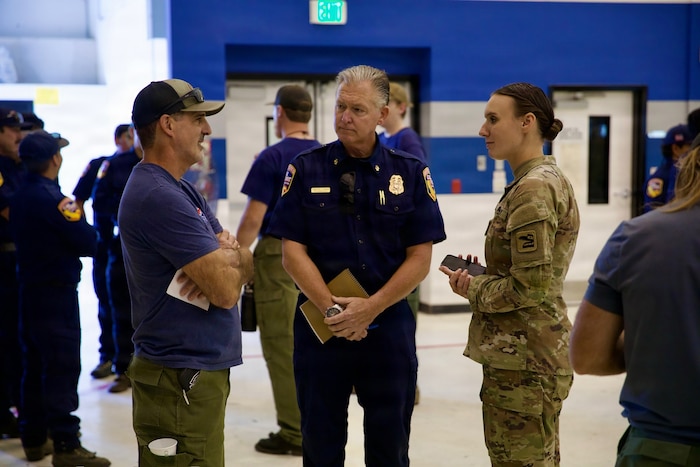 A man dressed in a police officer uniform talks to a man in a blue shirt and brown pants and a woman in a military camouflage uniform.