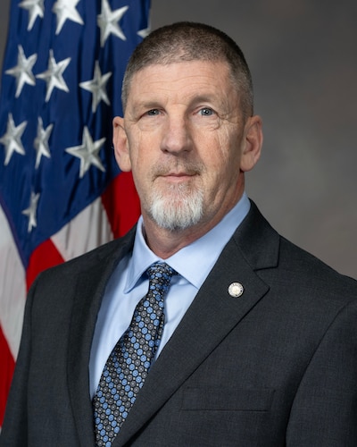 a man in a suit poses for an official photo in front of a US flag
