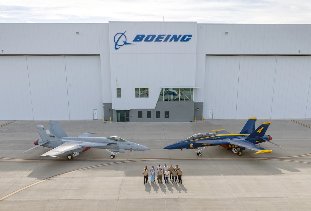 A group of people stands in front of an airplane hanger and between two fighter jets