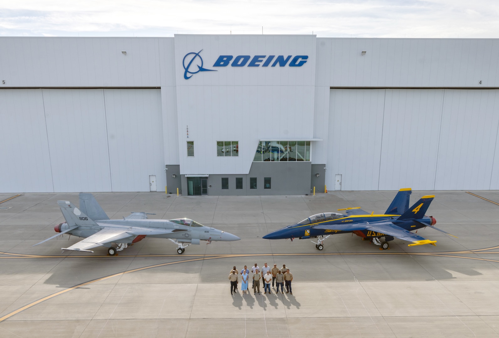 A group of people stands in front of an airplane hanger and between two fighter jets