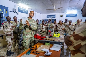 A military member points to a sheet of metal while instructing a room full of military students.