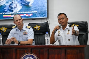 A man in a formal military uniform sits at a table and speaks into a microphone next to a man in similar attire.