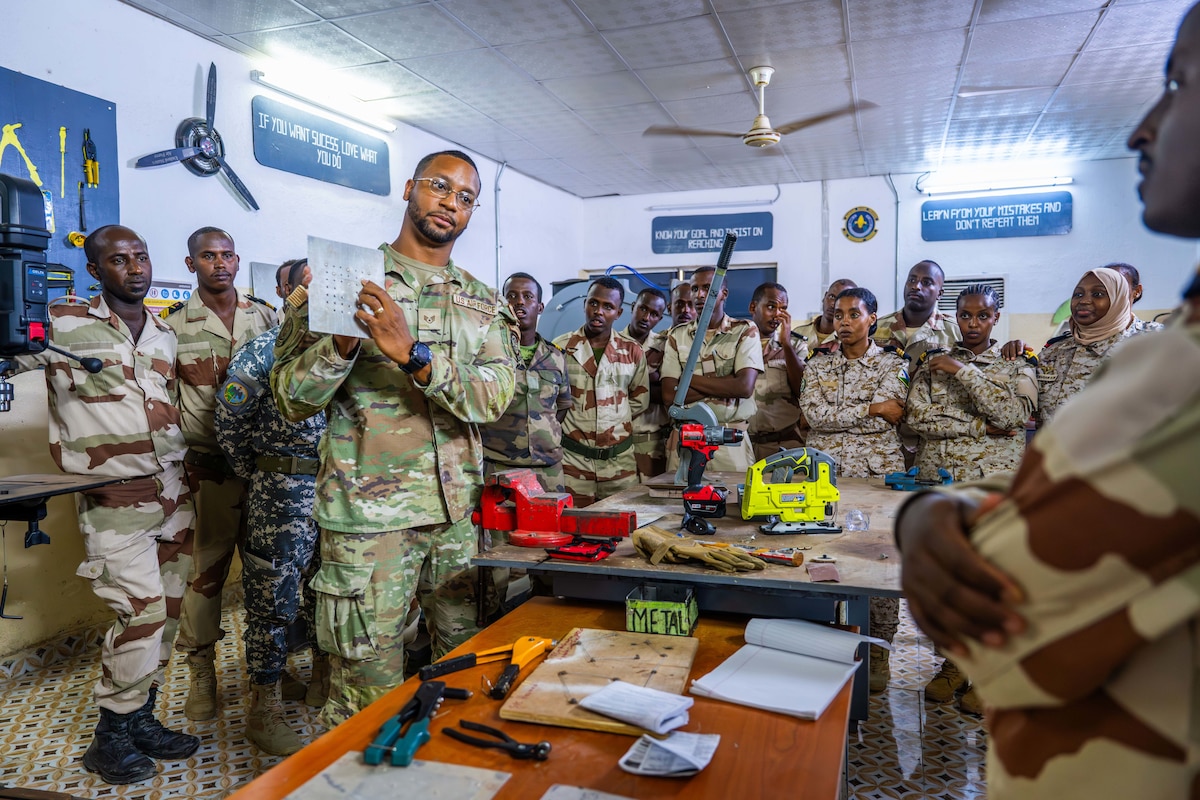 A military member points to a sheet of metal while instructing a room full of military students.