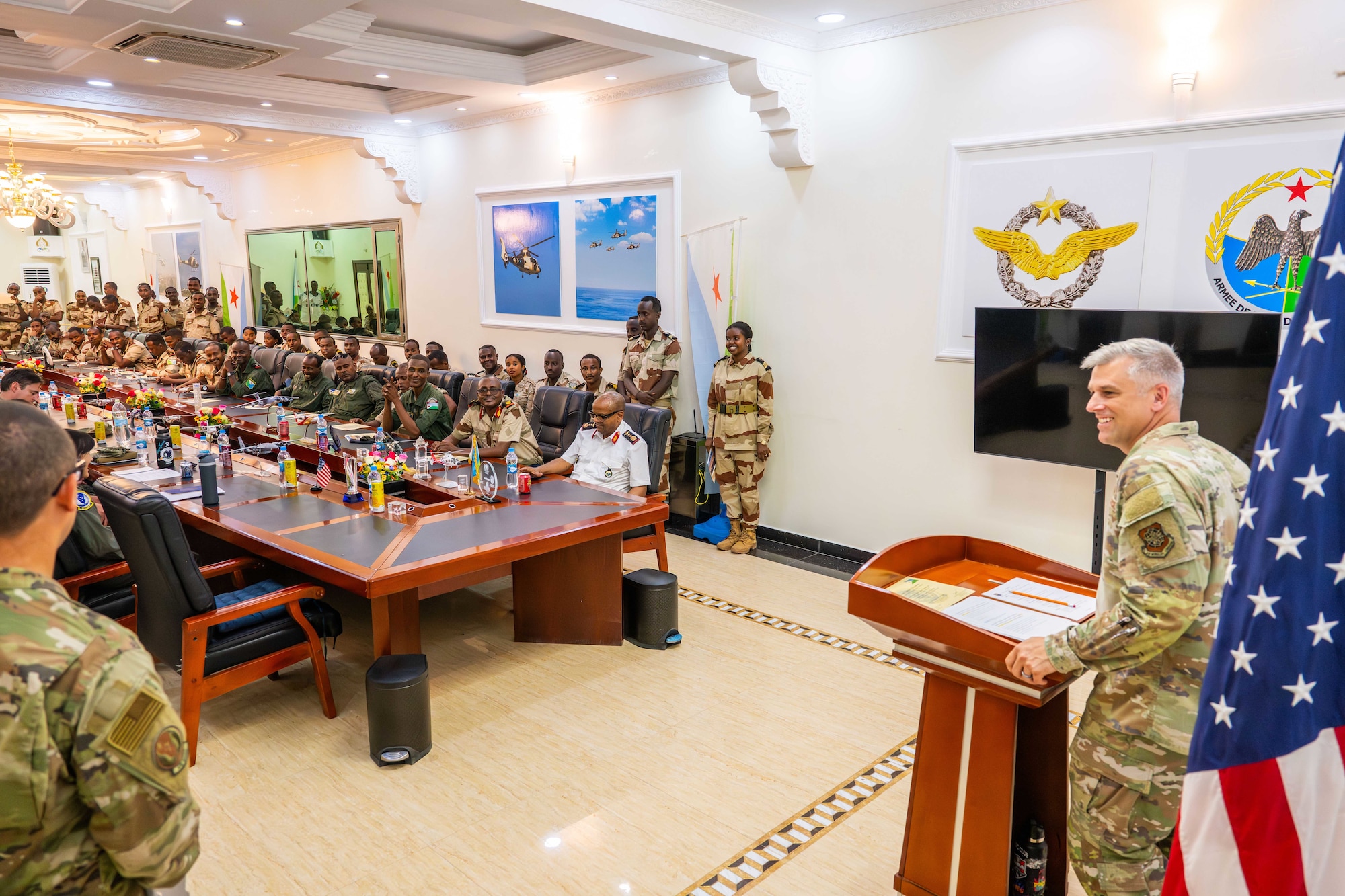 Military members sit in a room at a long table listening to a man speak.
