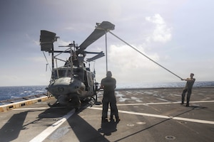 Three people wearing military uniforms use straps to maneuver the rotor blade of a helicopter parked on the deck of a military ship.
